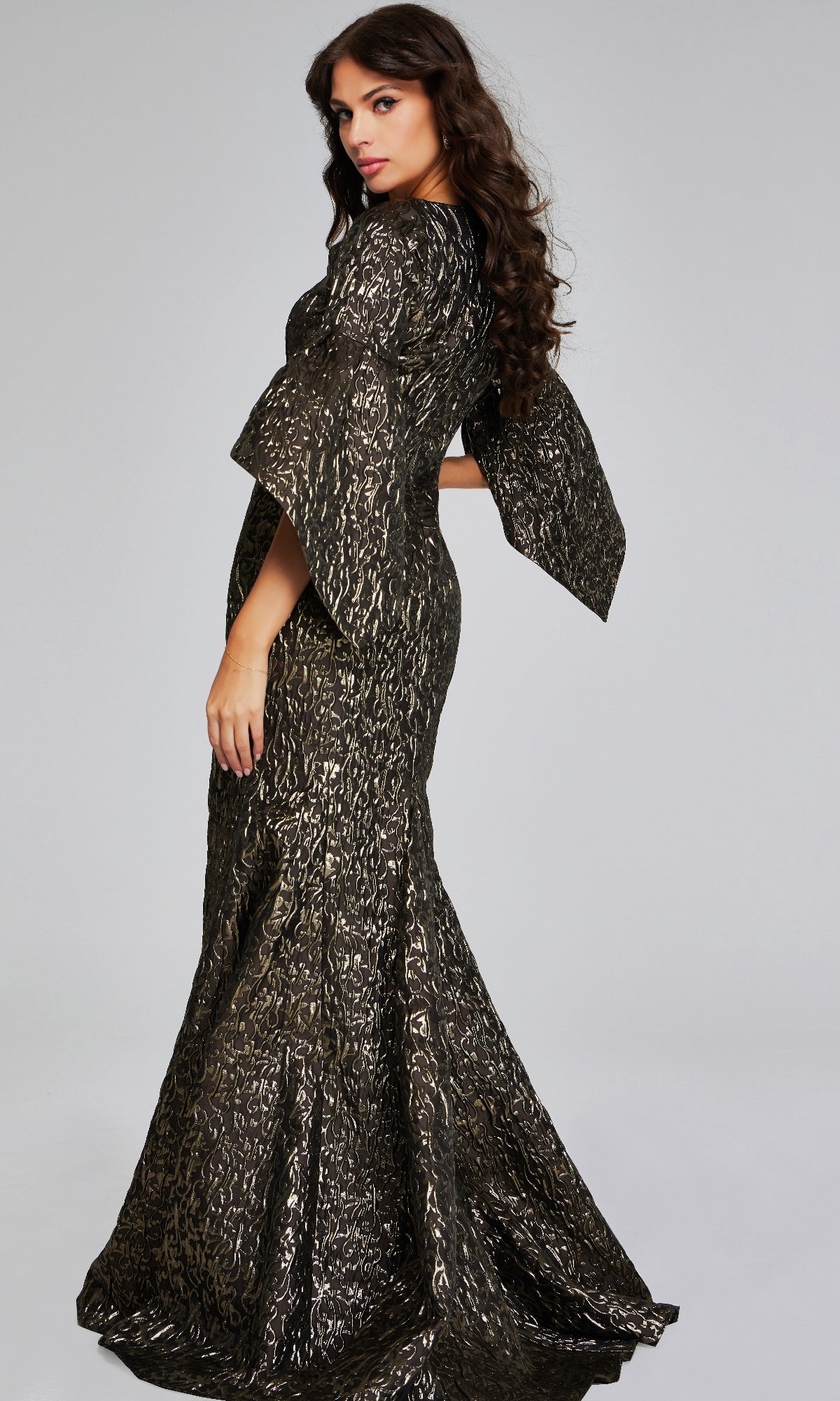 A woman poses in the Jovani 39080 long formal dress, featuring black and gold patterns and flared sleeves, against a plain gray background.